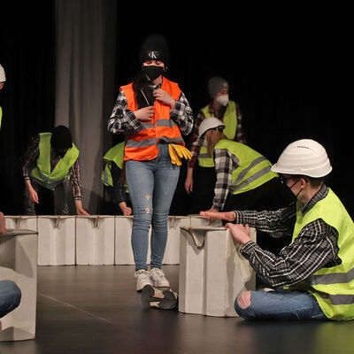 Auf einer Bühne spielen mehrere Schülerinnen und Schüler in gelben Warnwesten und Sturzhelmen die Bauphase nach und nutzen Pappkartons als Bausteine. Eine Person in der Mitte trägt eine orange Warnweste.