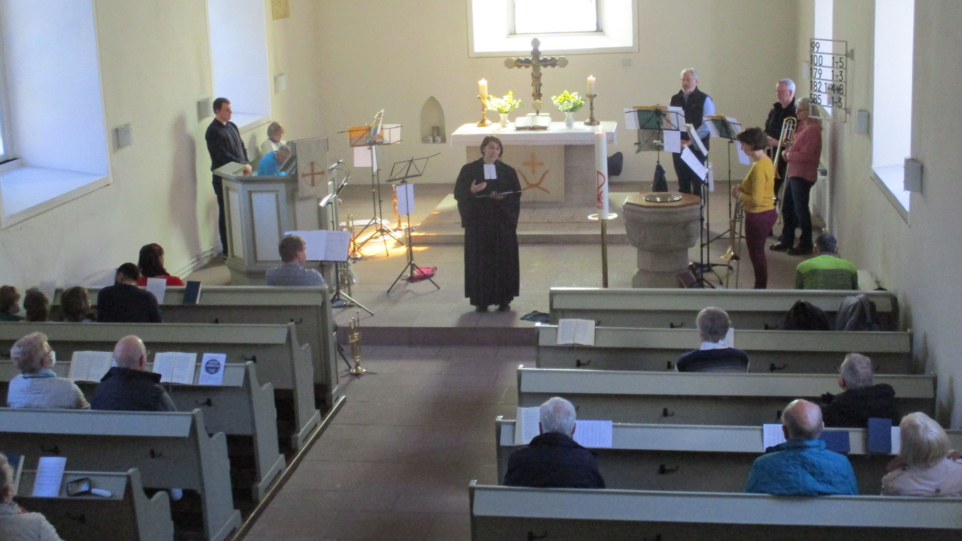 Blick von der Empore in den Kirchenraum: Die Pfarrerin predigt, der Posaunenchor steht im Altarraum, in den Reihen mit Holzbänken sitzen Bürgerinnen und Bürger.