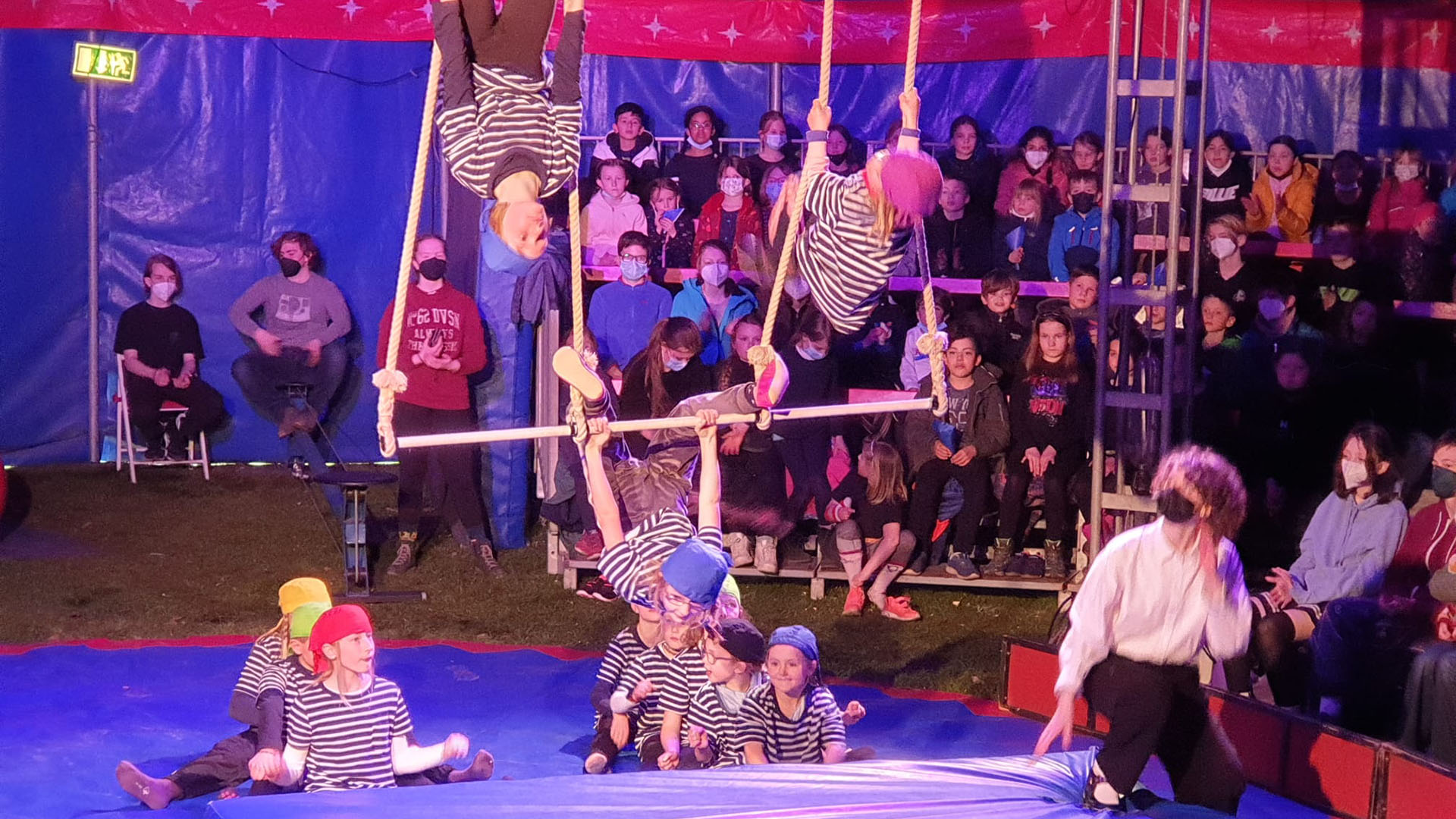 In einer Zirkusmanege turnen Kinder am Trapez, auf den Rängen dahinter sitzen Zuschauer.