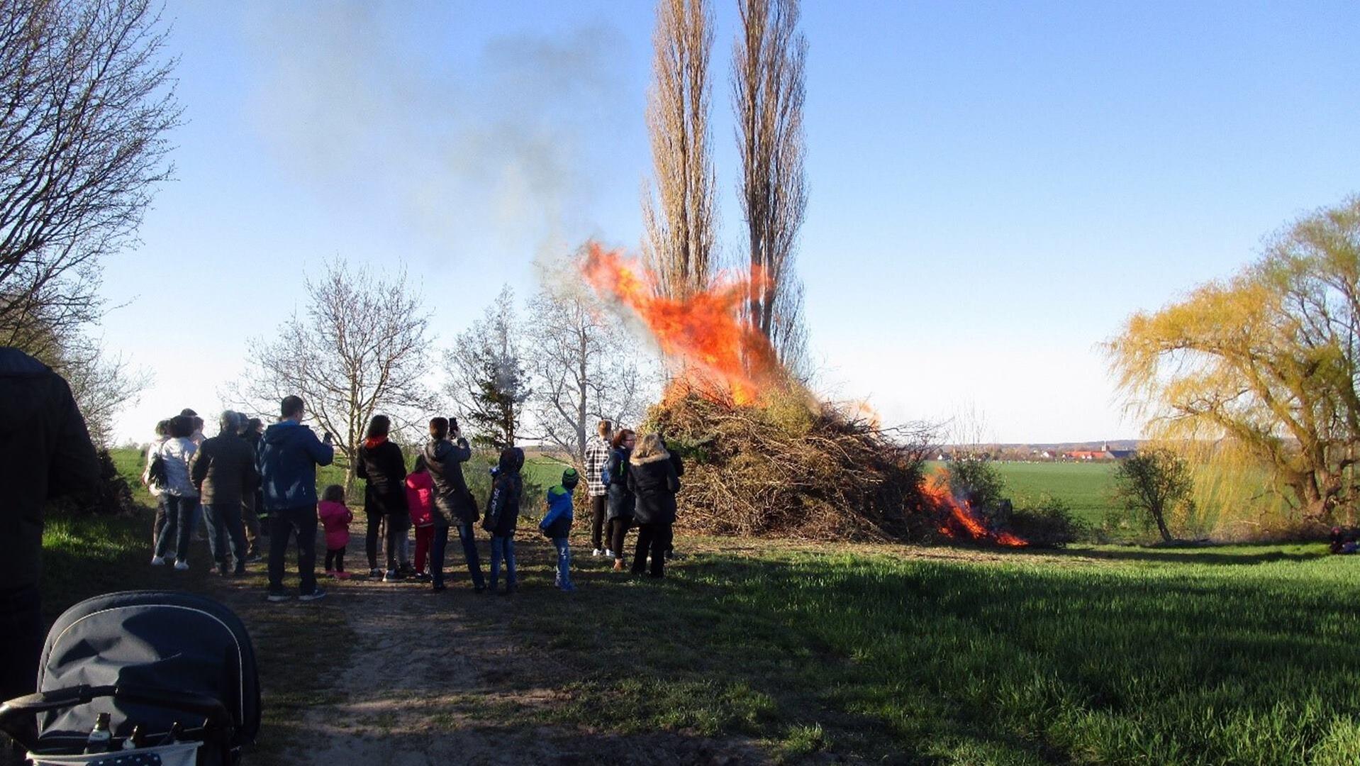 Ein brennender Haufen aufgestapelter Äste, davor steht eine große Personengruppe. Im Hintergrund stehen ein paar Bäume.