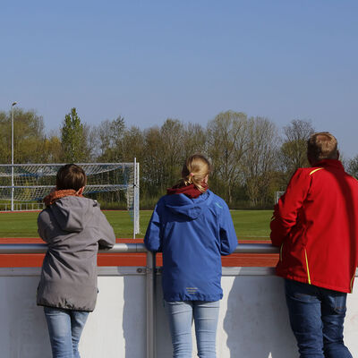 Ein Mann und drei Schüler stehen am Rande einer Sportanlage und schauen über die Bande auf die Sportfläche.