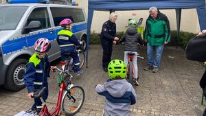 Einige Kinder mit Fahrrädern stehen in einer Reihe. Dahinter stehen ein Mann und eine Polizistin und überprüfen die Räder.