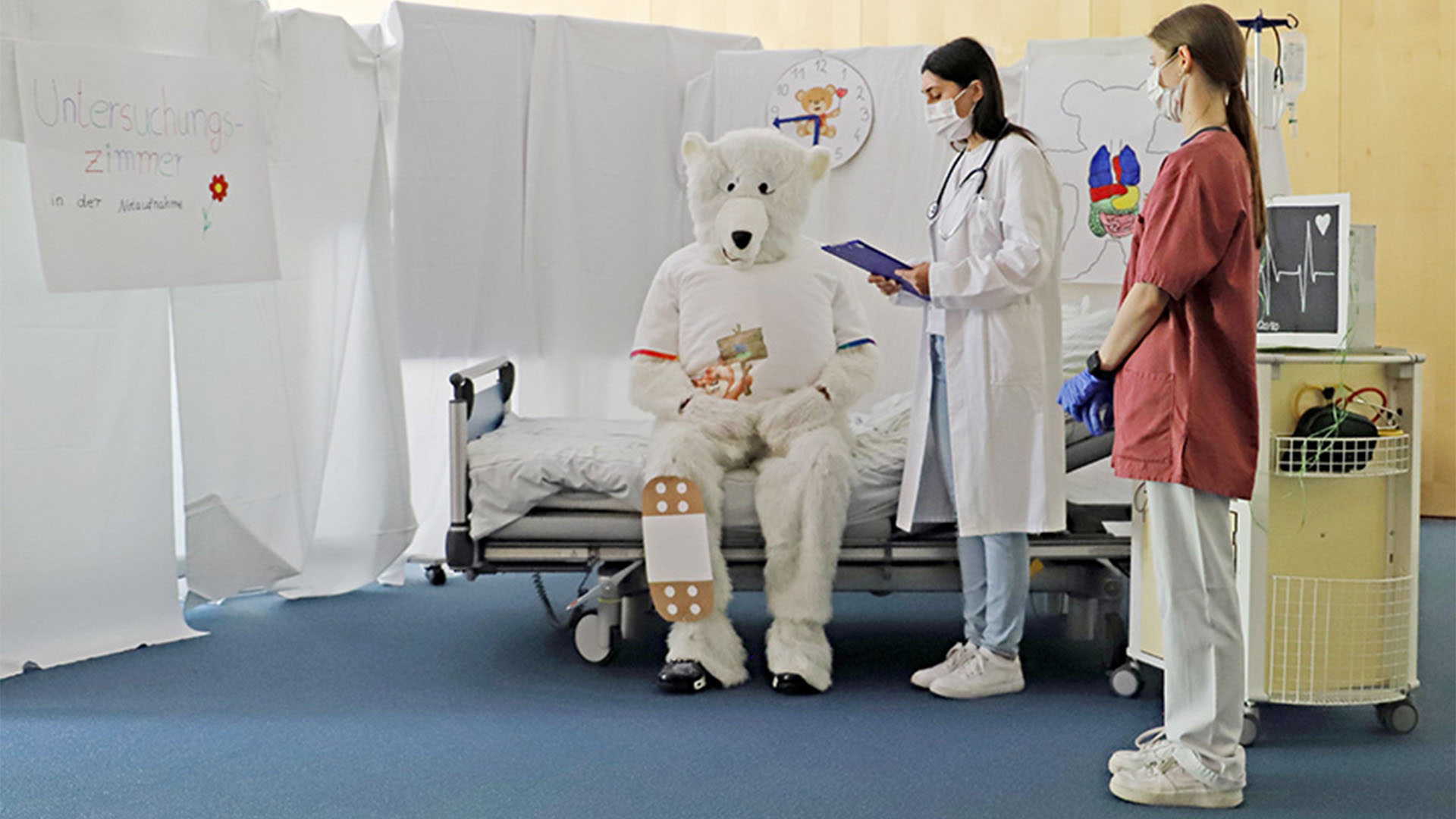 Ein Mensch im Bärenkostüm sitzt auf einem Krankenbett und hat ein großes Pflaster am Bein, eine Ärtzin steht daneben.