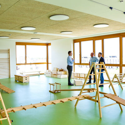 Sport- und Bewegungsraum mit verschiedenen Geräten aus Holz in einer Kindertagesstätte. Im Hintergrund stehen drei Männer, die sich an einer Fensterfront unterhalten.