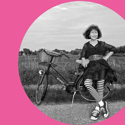 In einem runden Bildauschnitt lehnt sich eine Frau an einem Feldrand an ein abgestelltes Fahrrad. Außerhalb des Auschnittes befindet sich ein rosafarbener Rahmen mit der Beschriftung "Innitiatorinnen Bündnis 8. März"
