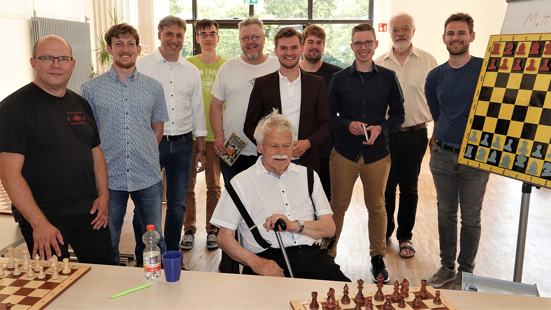 Ein älterer Mann sitzt vor einer Gruppe von zehn Männern und hat vor sich auf dem Tisch Schachbretter aufgebaut.