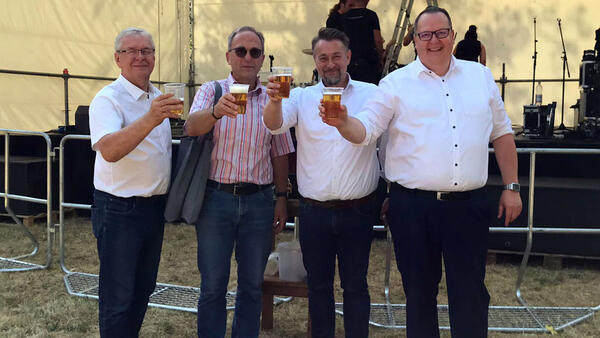 Gruppenfoto von vier Männern, die jeweilsmit einem Bierglas in der Hand zur Kamera prosten.