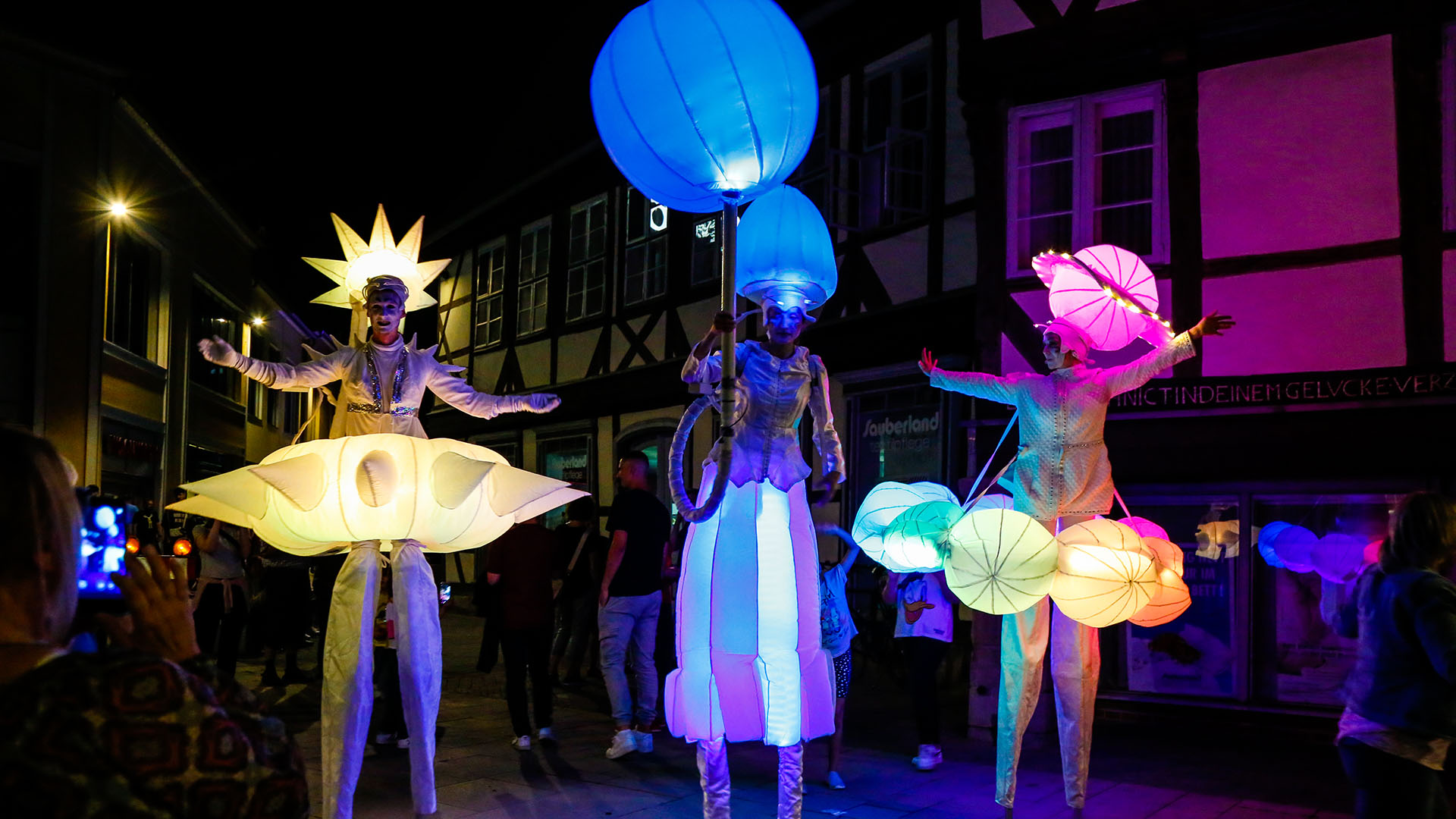 Drei Stelzenläufer mit bunt beleuchteten Fantasiekostümen laufen durch die Stadt
