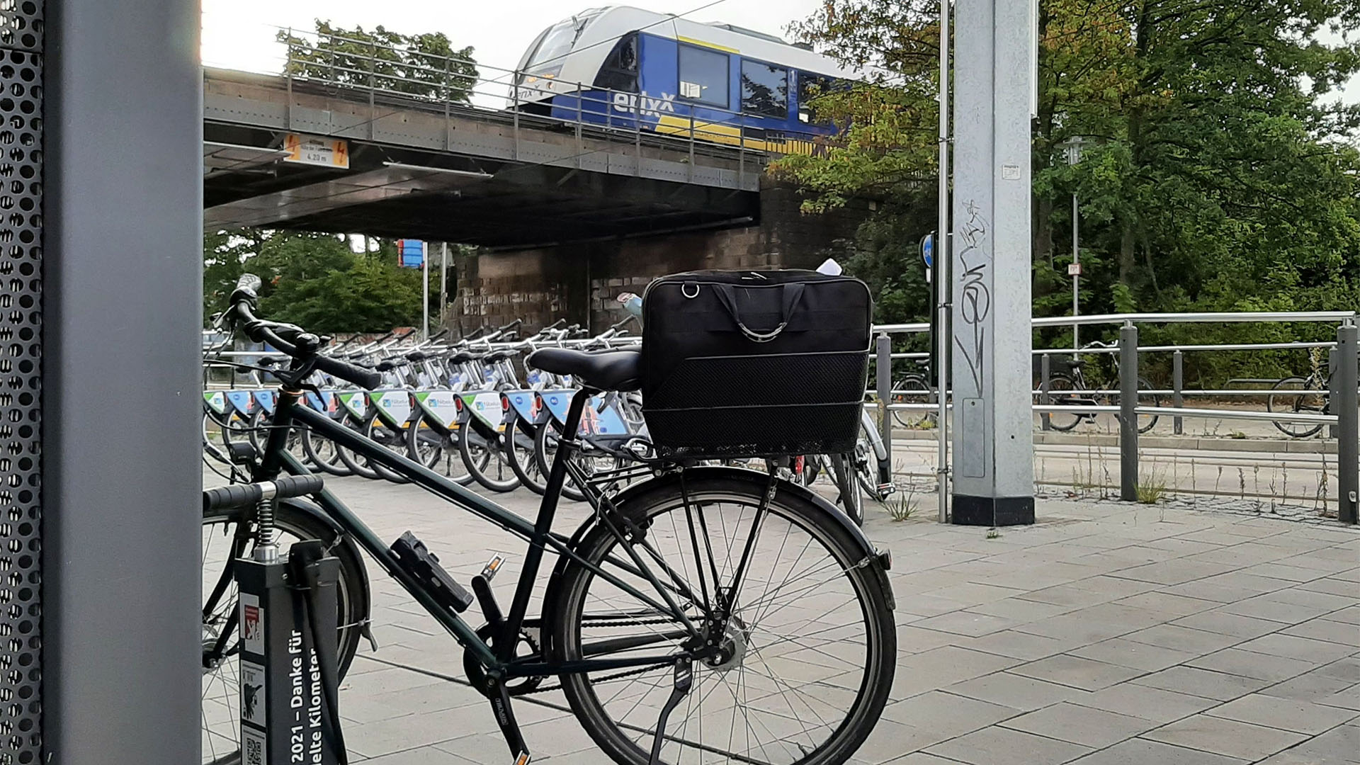 Ein Fahrrad steht an einem Fahrradständer, im Hintergrund eine Reihe von Leihfahrrädern an einer Straße. Über die Straße fürht eine Brücke, auf der gerade ein Zug fährt.