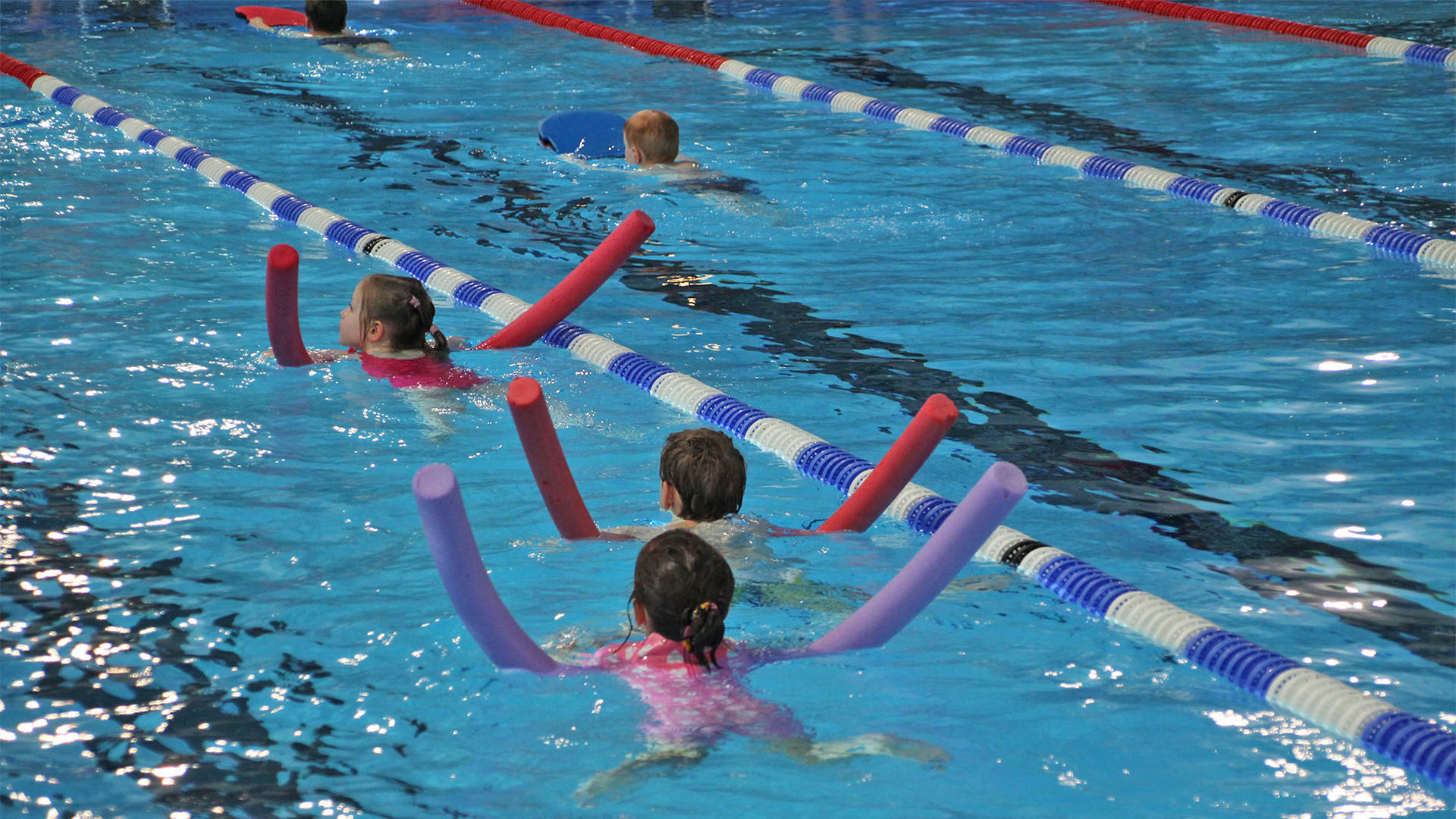 Kinder schwimmen in einem Schwimmbecken hintereinander. Jedes Kind hat eine farbige Schwimmnudel unter dem Körper.