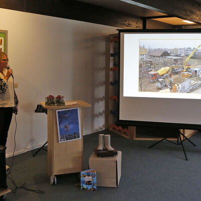 Eine Frau steht mit einem Miktrofon in der Hand neben einer Leinwand, auf der ein Foto einer Baustelle mit Baufahrzeugen zu sehen ist.