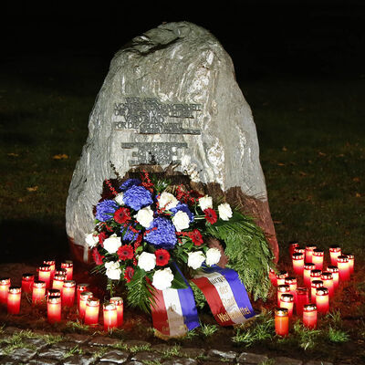 Ein einem Gedenkstein lehnt ein Kranz mit Schleife, davor stehen brennende Kerzenlichter.