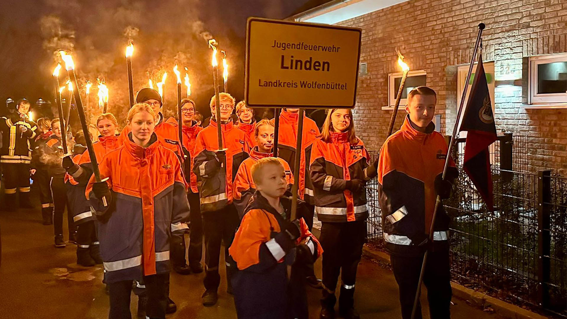 Jugendliche der Jugendfeuerwehr Linden führen mit Fackeln in den Händen und in Uniform den Laternenumzug an. Ein Junge trägt ein Schild "Jugendfeuerwehr Linden, Landkreis Wolfenbüttel", ein anderer eine Fahne.