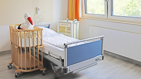 Blick in ein Zimmer im Krankenhaus, neben dem Krankenbett steht ein Stubenwagen aus Holz, in dem ein Stoff-Storch sitzt.