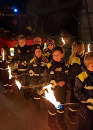Mehrere Kinder in Feuerwehrkleidung stehen hintereinander mit brennenden Fackeln in den Händen.