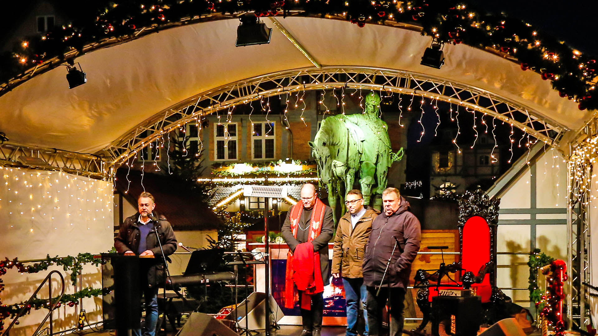 Vier Männer stehen auf einer weihnachtlich geschmückten Bühne, einer davon am Mikrofon. Im Hintergrund erkennt man das beleuchtete Standbild auf dem Stadtmarkt und Weihnachtsbuden des Weihnachtsmarktes.