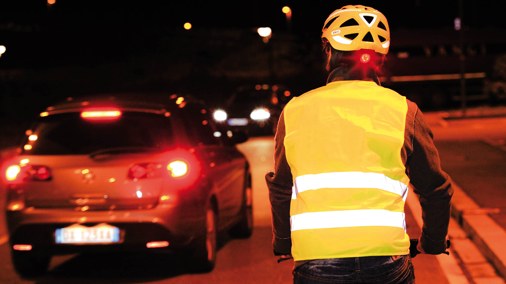 Ein Radfahrer fährt mit gelber Warnweste und Helm, beides mit Reflektorstreifen, im Dunklen im Straßenverkehr.