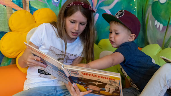 Ein  Mädchen und ein kleinerer Junge lesen gemeinsam in der Stadtbücherei in einem Buch.