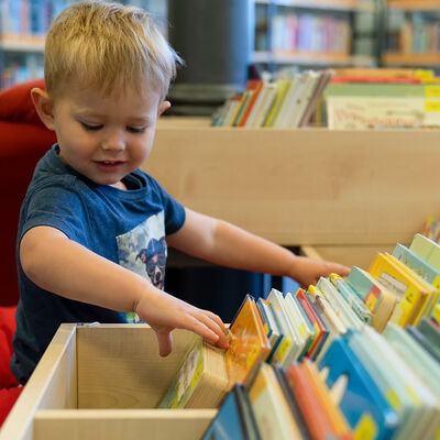 Ein Junge schaut in der Stadtbücherei durch eine Sammlung von Kinderbüchern, die in einer Holzkiste aufgestellt sind