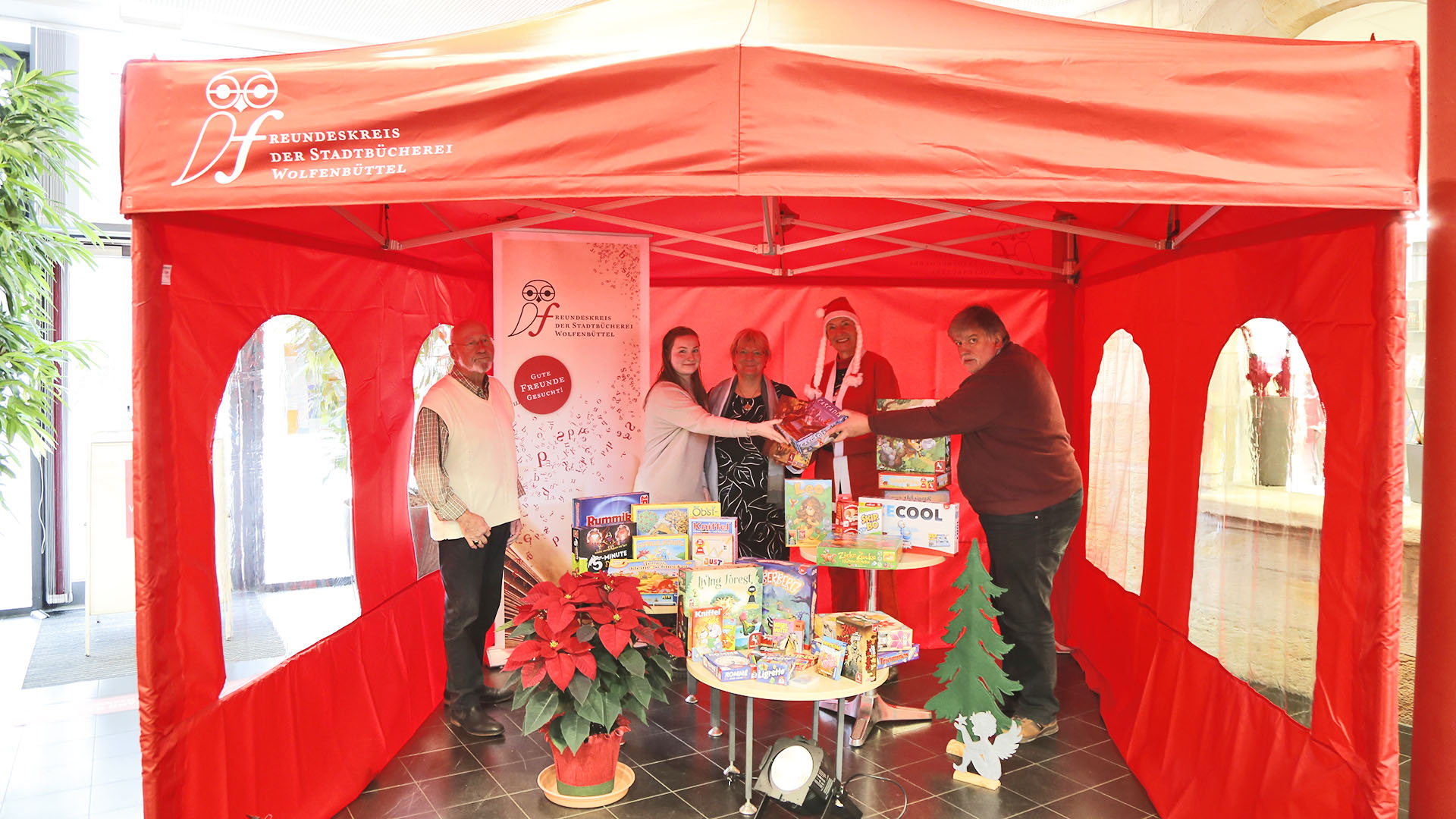 Zwei Männer und drei Frauen, eine davon im Weihnachtsmannkostüm, stehen in einem roten Pavillon. Vor ihnen Tischchen mit Gesellschaftsspielen.