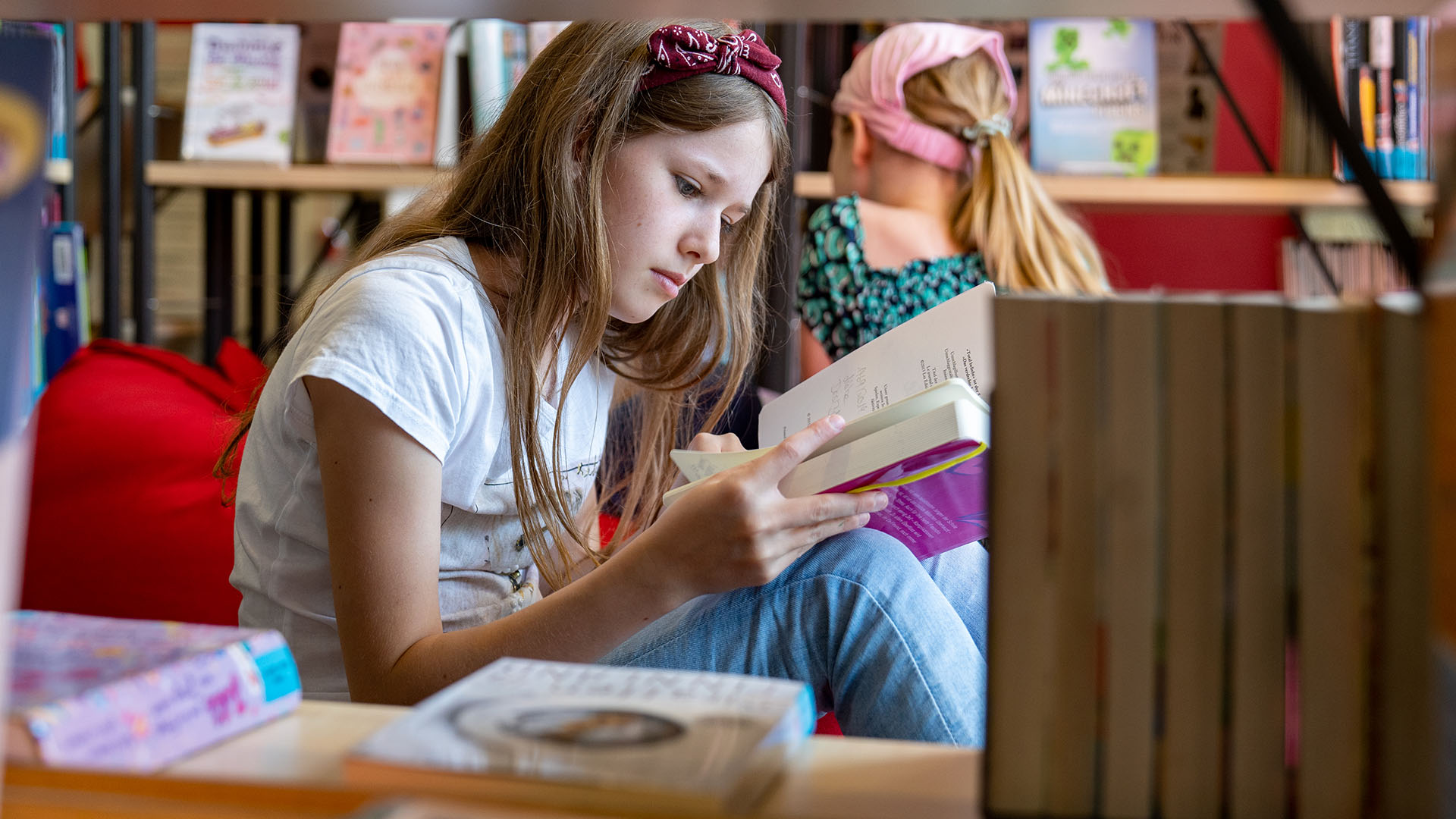 Ein Mädchen sitzt in der Stadtbücherei und liest in einem Buch, im Hintergrund schaut ein anderes Mädchen Bücher im Bücherregal an.