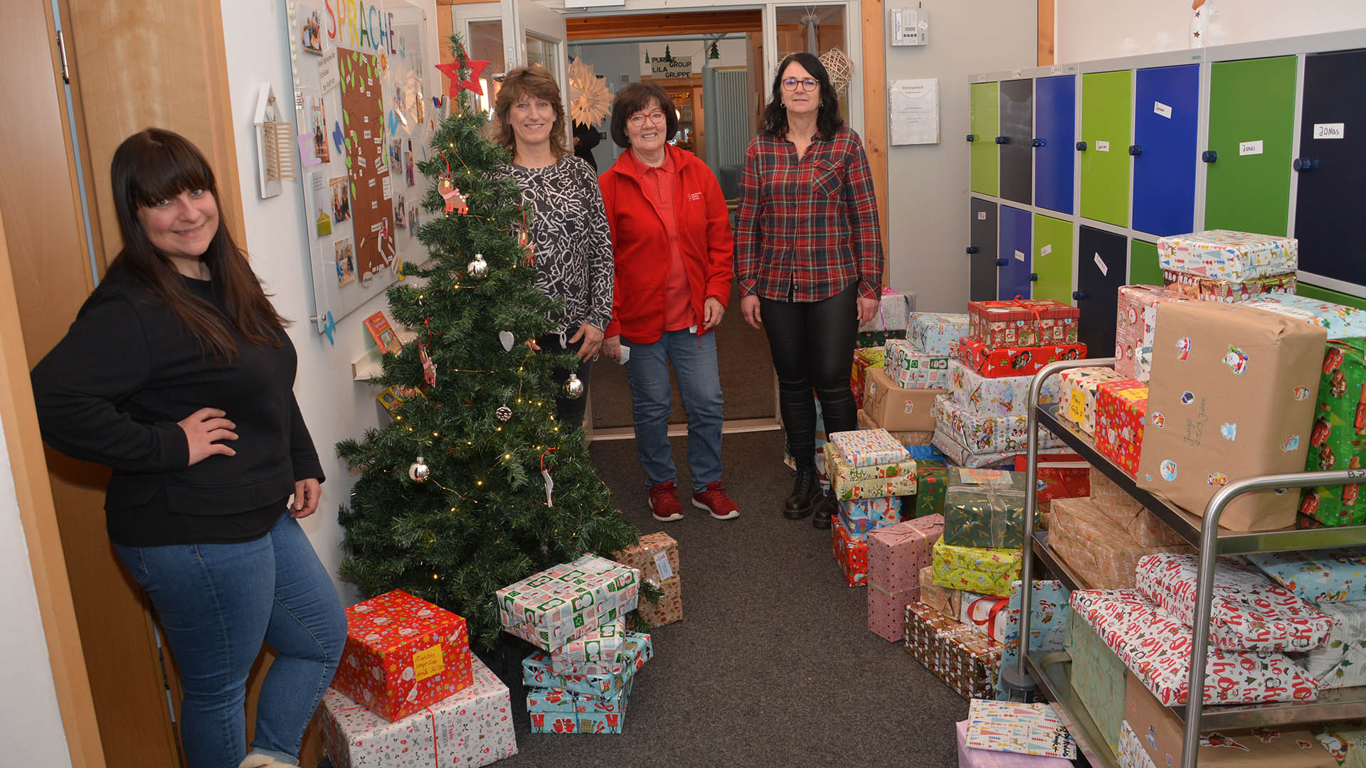 Vier Frauen stehen neben einem Weihnachtsbaum und vielen Stapeln von weihnachtlich verpackten Geschenken.
