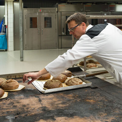 Ein Bäcker in der Backstube arbeitet an einem Tisch mit mehreren Blecken mit Brot, ein junger Lehrling schaut ihm zu.