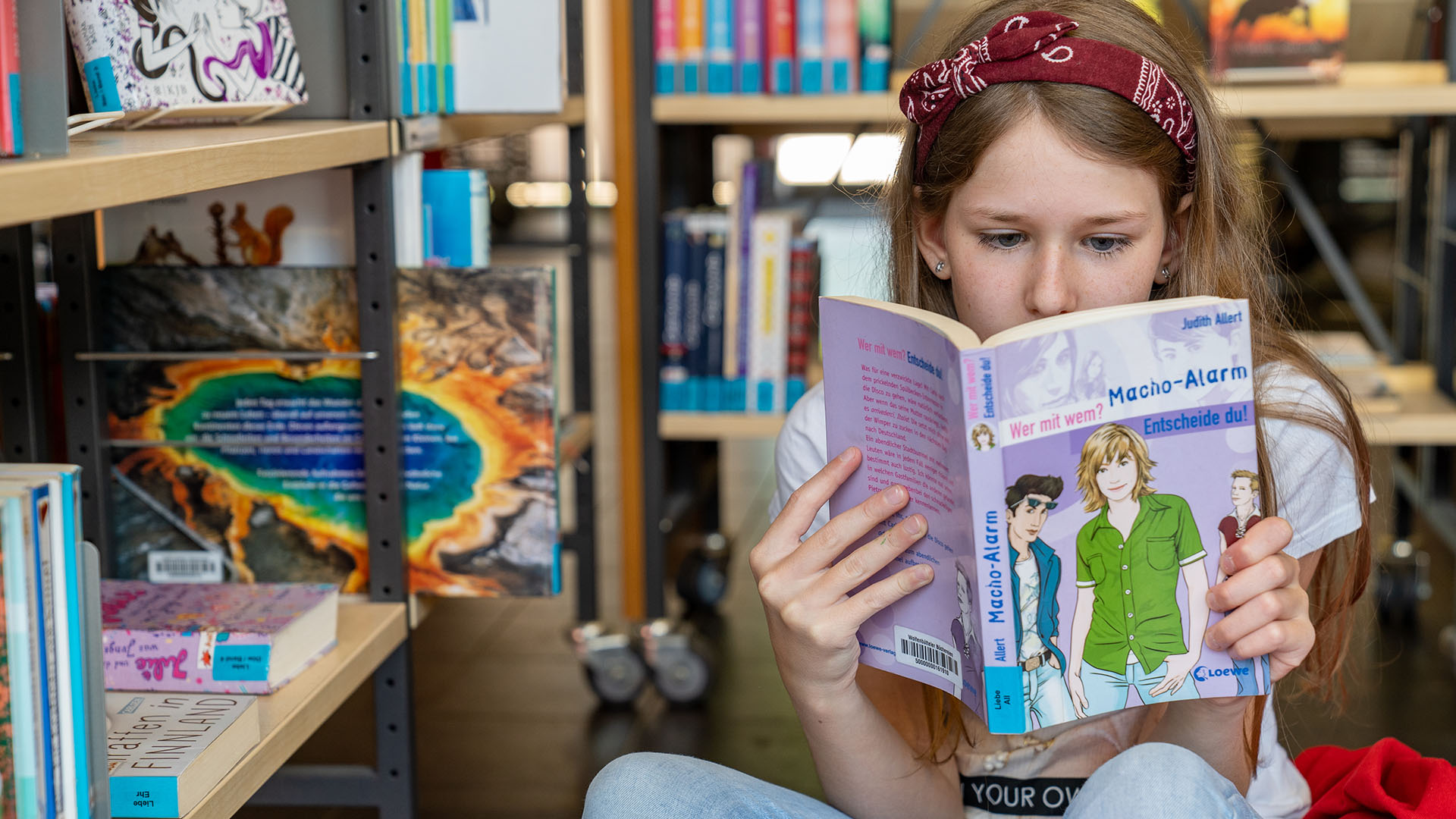Ein Mädchen sitzt in der Stadtbücherei und liest in einem Buch, hinter und neben ihr stehen Buchregale