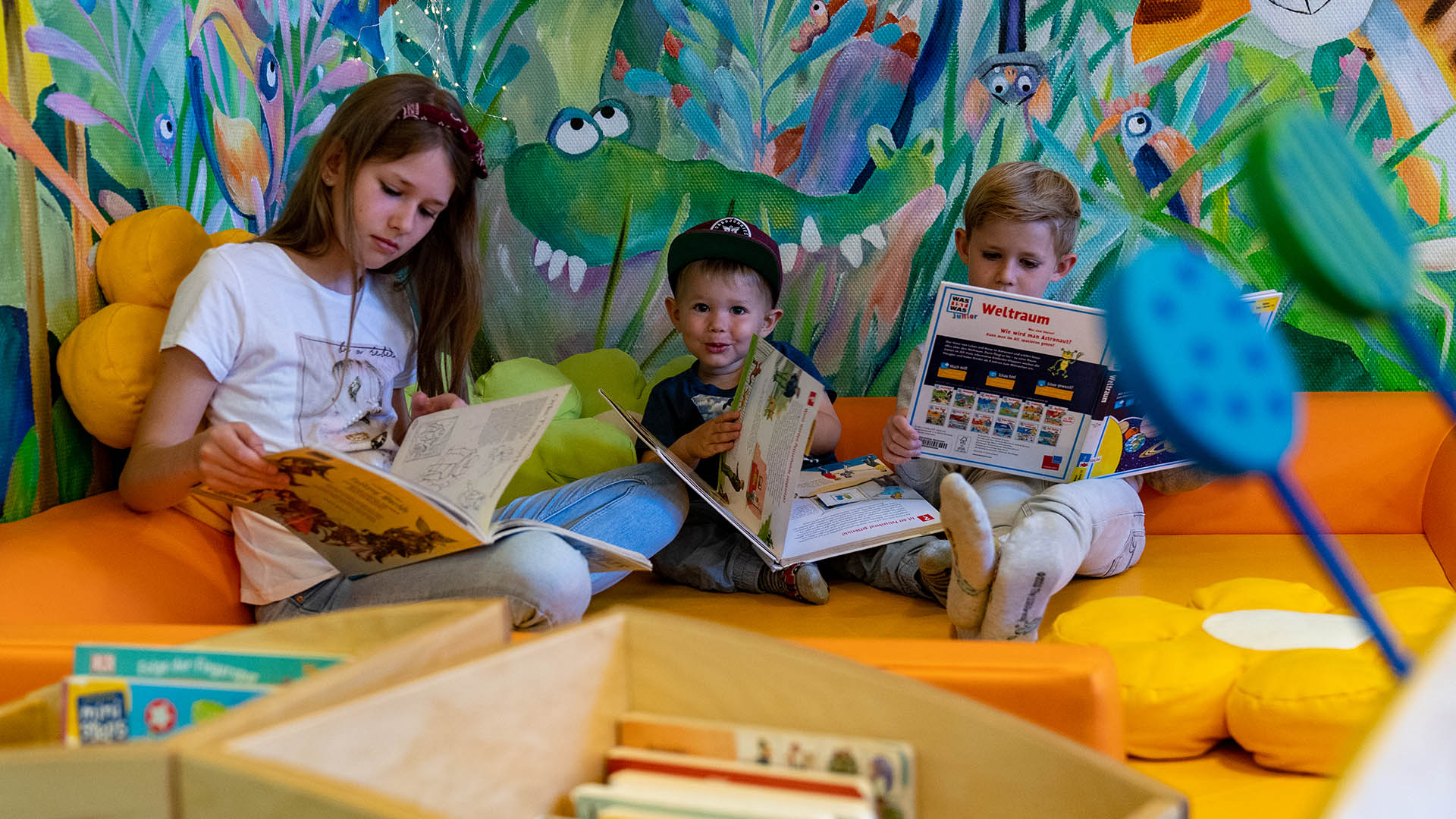 Ein Mädchen und zwei Jungen sitzen in der Stadtbücherei vor einer Wand, die mit Dschungelmotiven bemalt ist, und lesen in Kinder- und Jugendbüchern.