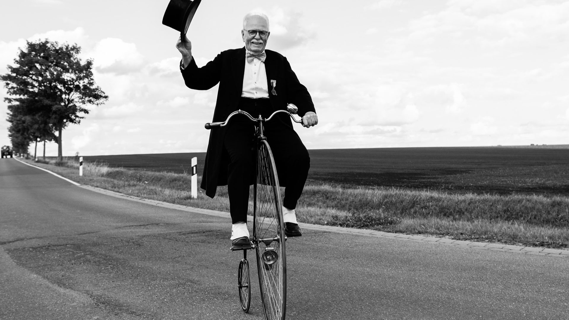 Schwarzweißfots eines Mannes, der in Smoking und Zylinder, mit Gamaschen und Fliege auf einem Hochrad sitzt und über eine Straße fährt. Den Zylinder schwenkt er.