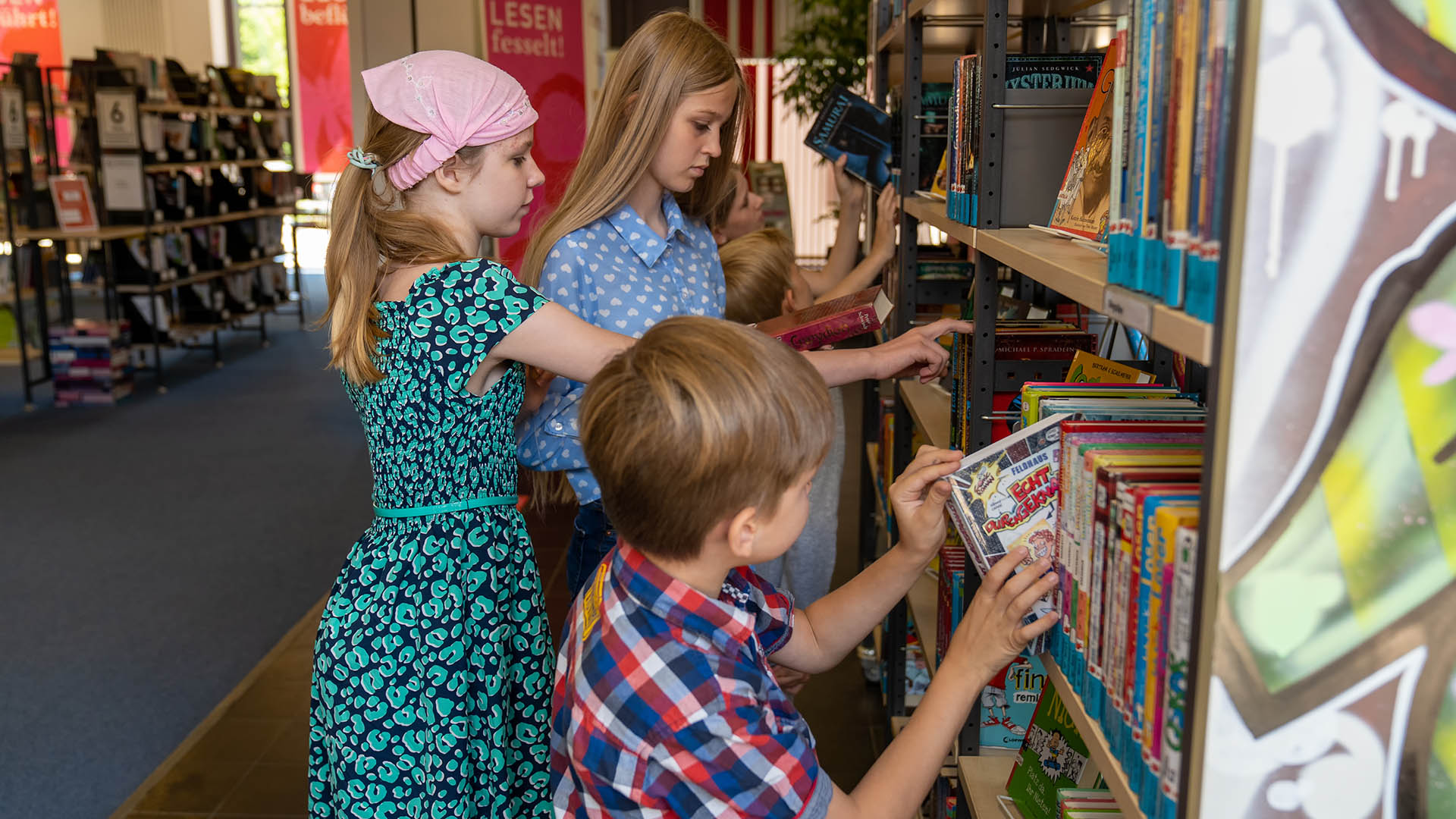 Zwei Mädchen und ein Junge stehen in der Stadtbücherei vor einem Bücherregal und suchen sich Bücher aus