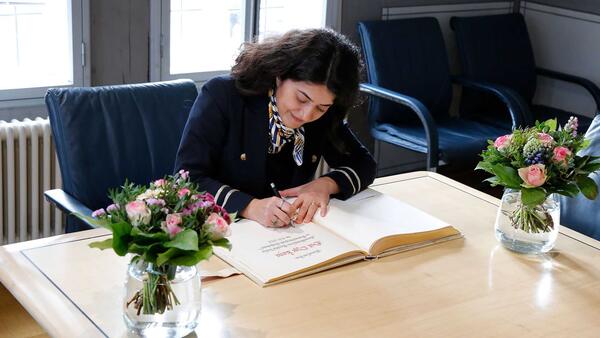 Eine Frau sitzt an einem mit zwei Blumensträußen geschmückten Tisch und schreibt in ein dickes Buch.