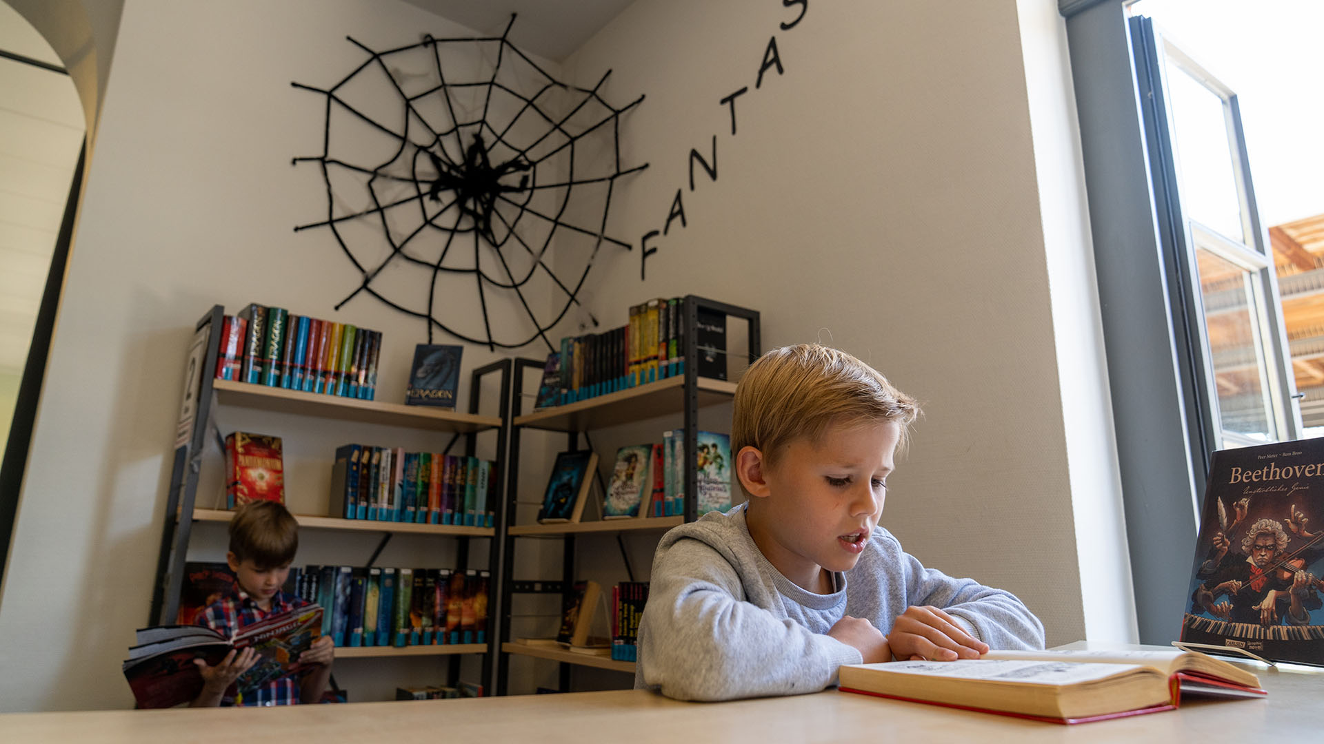 Ein kleinerer Junge liest in der Stadtbücherei in einem Buch, hinter ihm hängt über einem Bücherregal ein großes schwarzes Spinnennetz mit dem Schriftzug Fantasy