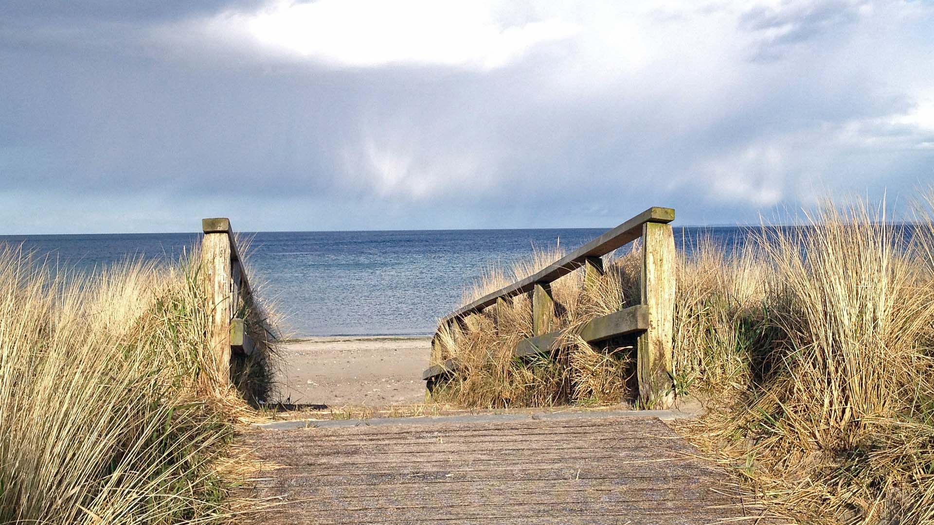 Ein hölzerner Bohlenweg mündet in einer Teppe, die zum Ostseestrand führt. Rechts und links neben der Treppe Holzgeländer und Standhafer. Über dem Meer sind weiße Wolken.
