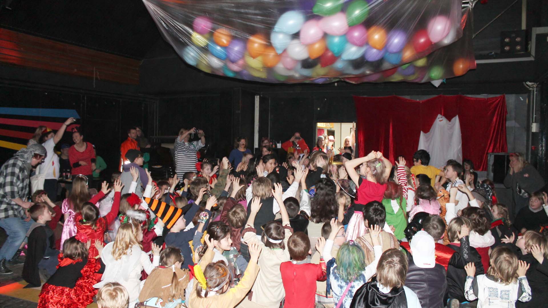 Viele Kinder greifen nach Luftballons, die von der Decke in einer Folie hängen.