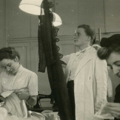 historisches Schwarzweißfoto: vier Schneiderinnen sitzen oder stehen neben einem Nätisch und arbeiten an Kleidungsstücken