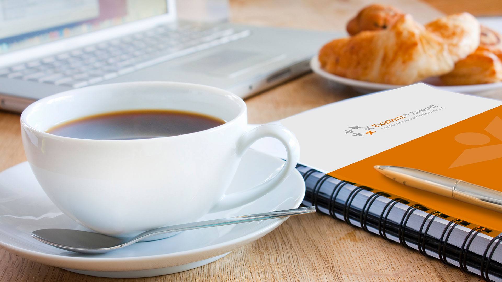 Auf einem Tisch steht eine Tasse Kaffee, ein Laptop und ein Teller mit Gebäck, dazwischen liegt ein Spiralblock mit dem Logo des Vereins Existenz&Zukunft und ein Stift