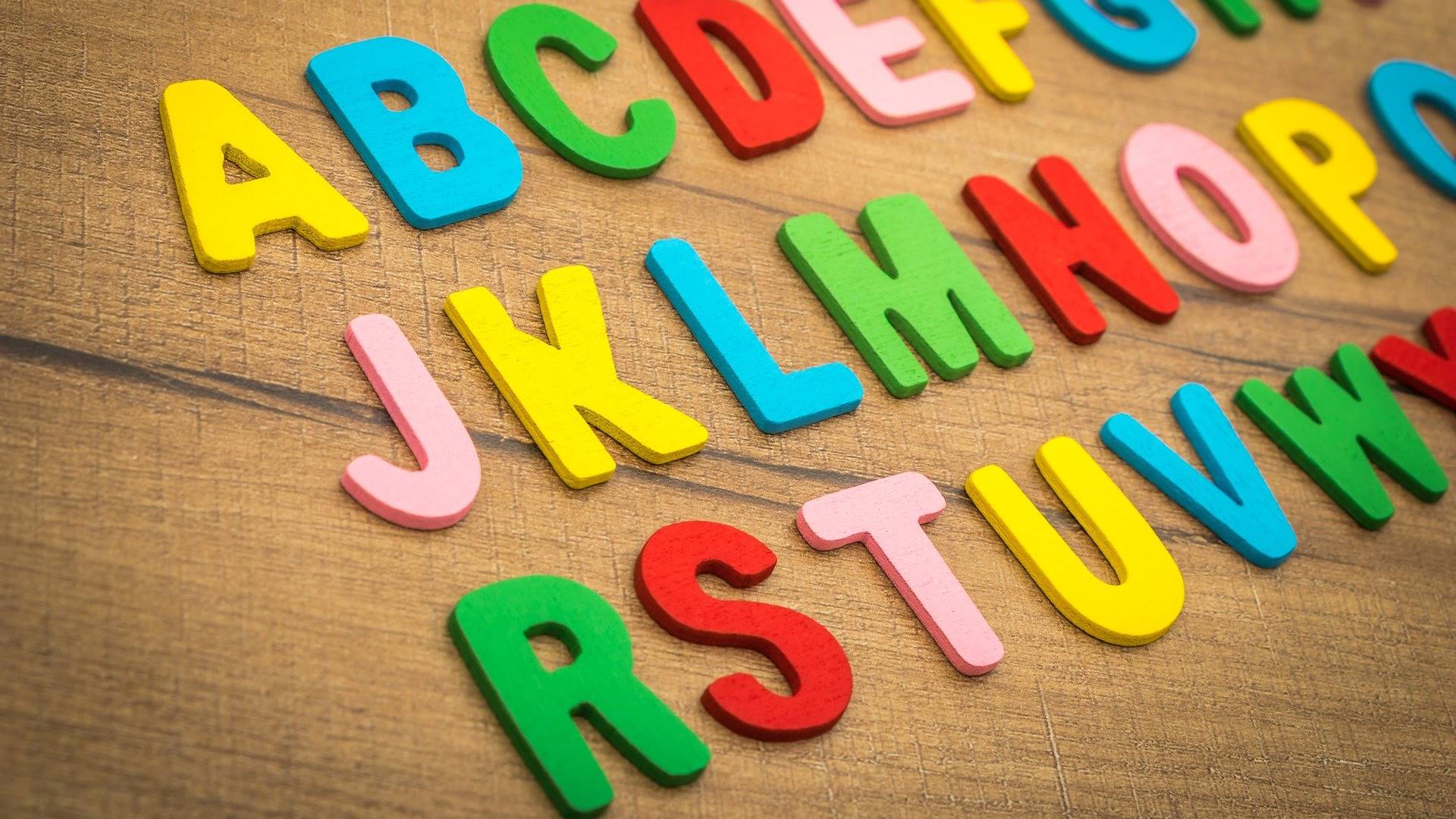 Hölzerne bunte Buchstaben sind alphabetisch in drei Zeilen auf einem Stück Holz aufgereiht. Die Buchstaben sind Gelb, Blau, Grün, Rot und Rosa gefärbt.