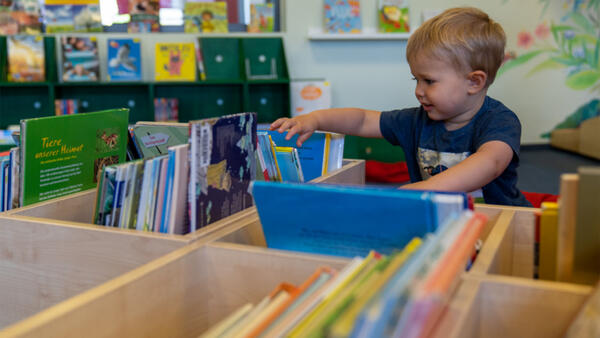 Ein Junge schaut in der Stadtbücherei durch eine Sammlung von Kinderbüchern, die in einer Holzkiste aufgestellt sind