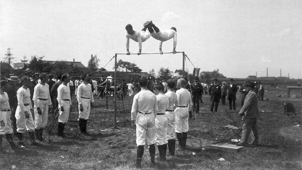 Schwarzweißfoto eines Turnfest des MTV Wolfenbüttel 1910, zwei Männer schwingen am Hochreck, andere schauen zu.
