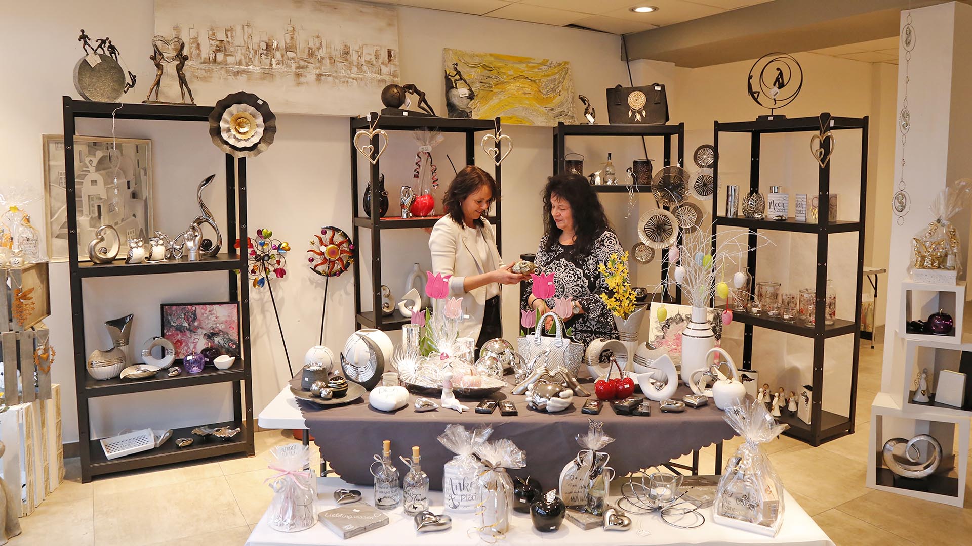 Zwei Frauen stehenin einem Geschäft mit Deko- und Einrichtungsgegenständen, die auf zwei Tischen und in Regalen präsentiert werden. Beide schauen sich ein Dekoobjekt an.