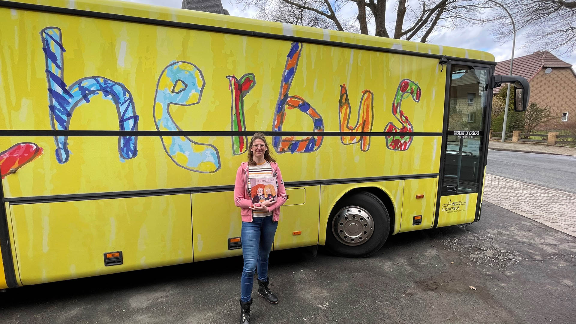 Eine Frau steht mit einem Buch in den Händen vor einem gelben Bus, auf dem noch ein Teil des Wortes "Bücherbus" zu lesen ist