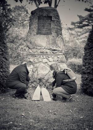 Ein Mann und eine Frau legen an einem Gedenkstein einen Kranz mit zwei breiten Bndern nieder.