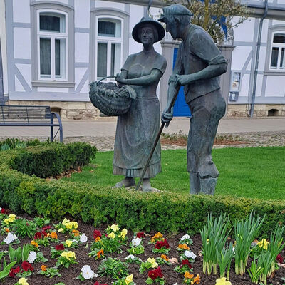 Zwei Statuen von einer Frau und einem Mann auf einer  Rasenfläche, davor ein beet mit Stiefmütterchen und Frühblühern