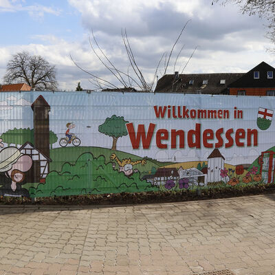 An einem Zauni st ein langes Banner befestigt mit dem Schriftzug "Willkommen in Wendessen". Daneben sind in Comicform Figuren und Motive aus Wendessen zu sehen.