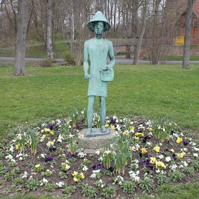 Eine Statue steht auf einer Rasenfläche. In einem Kreis um die Statue sind Blumen gepflanzt.