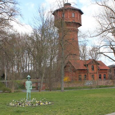 Auf einer Rasenfläche steht eine Statue von einem kreisrunden Blumenbeet umgeben. Im Hintergrund ist ein hoher Turm zu erkennen.