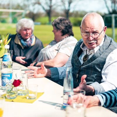 An einem mit Blumen und Gläsern gedeckten Tisch diskutiert ein älterer Mann mit drei Frauen.