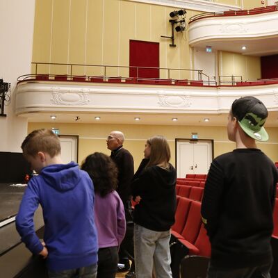 In einem großen Theatersaal stehen vier Jugendliche, ein Mann und eine Frau vor einer Bühne.