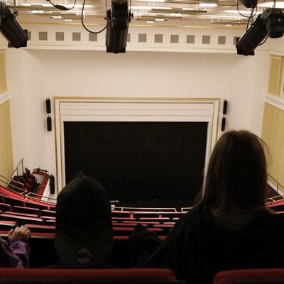 In einem leeren Theatersaal sitzen drei Kinder auf dem erhöhten zweiten Rang und blicken auf die leere Bühne.